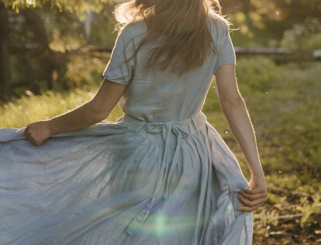 A woman facing away from the camera, holding her skirt while walking on dewy grass at sunrise. The image conveys a feeling of freedom and serenity.