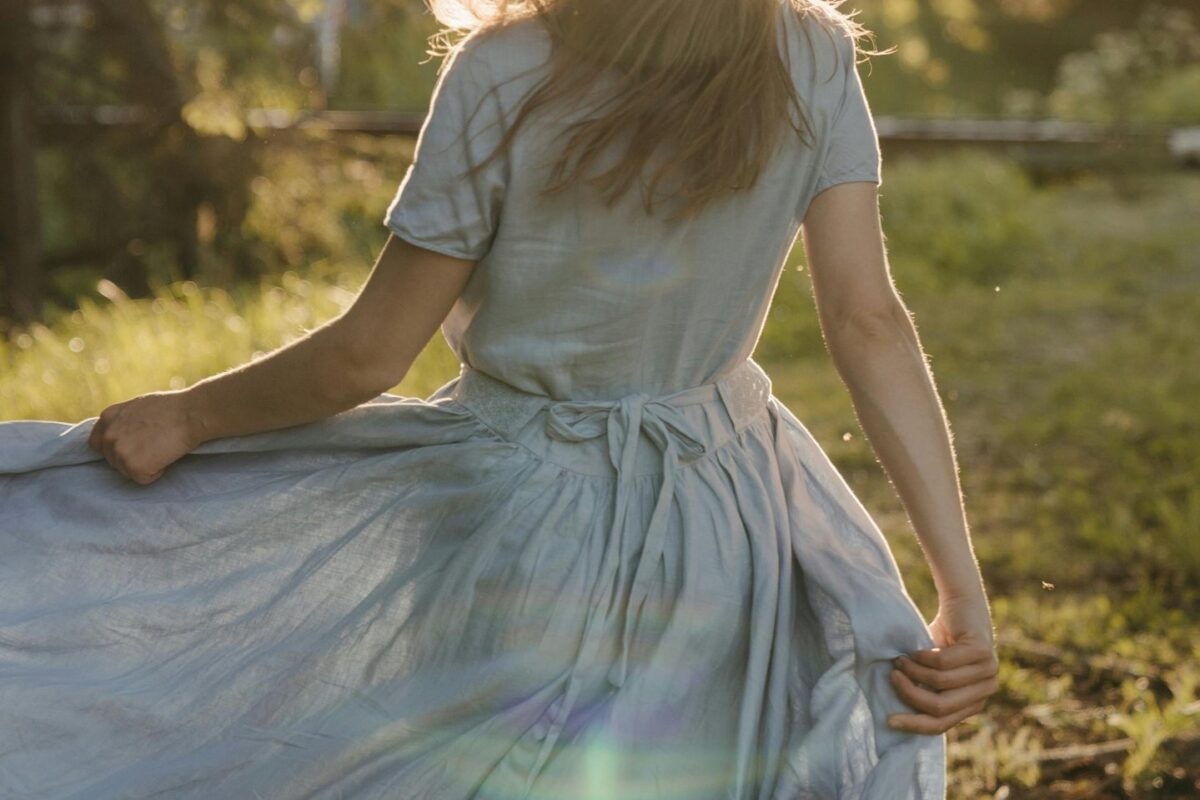 A woman facing away from the camera, holding her skirt while walking on dewy grass at sunrise. The image conveys a feeling of freedom and serenity.