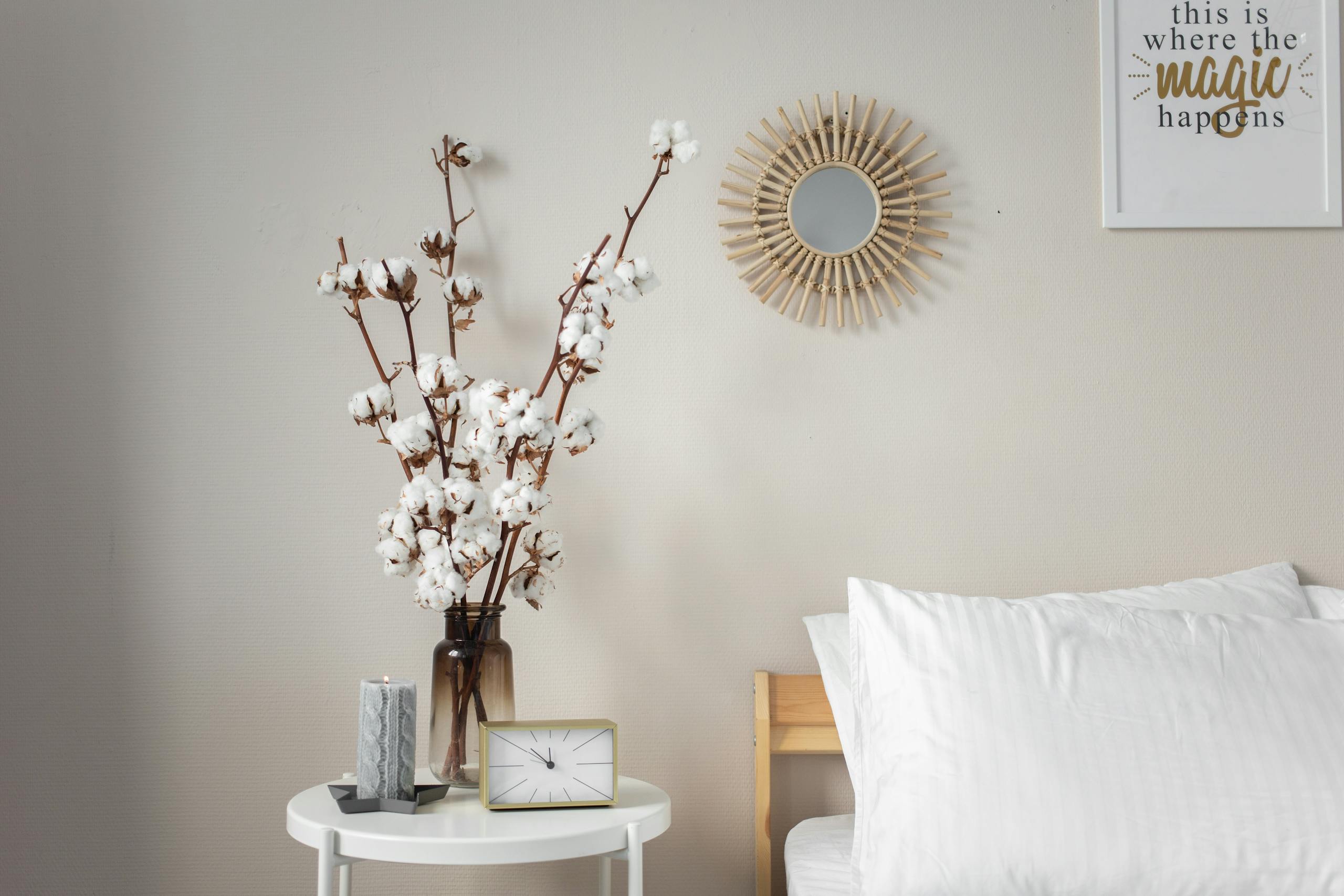 A calming bedroom scene, minimalist bedside table with cotton flowers, candle, table clock and modern decor details like a mirror and wall art — calm home decor for peaceful mornings.