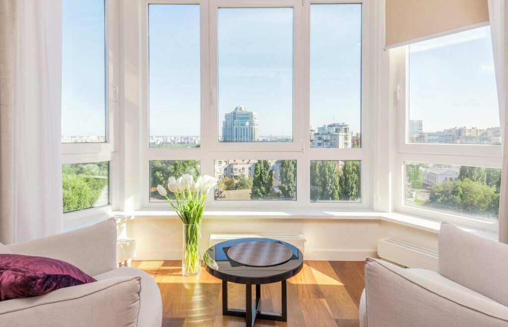 Sunlit minimalist living space reflecting calm and balance. Interior of modern penthouse with comfortable white armchairs and round side table placed near windows overlooking city in sunny day