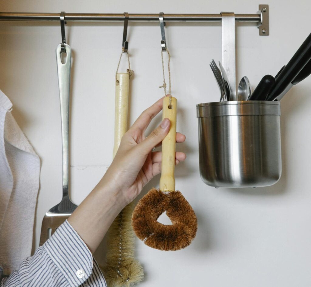 Simple, decluttered kitchen utensils showing minimalist home wellness and mindful living. A female hanging brush for washing dishes on metal rack on wall with various equipment in light kitchen at home.