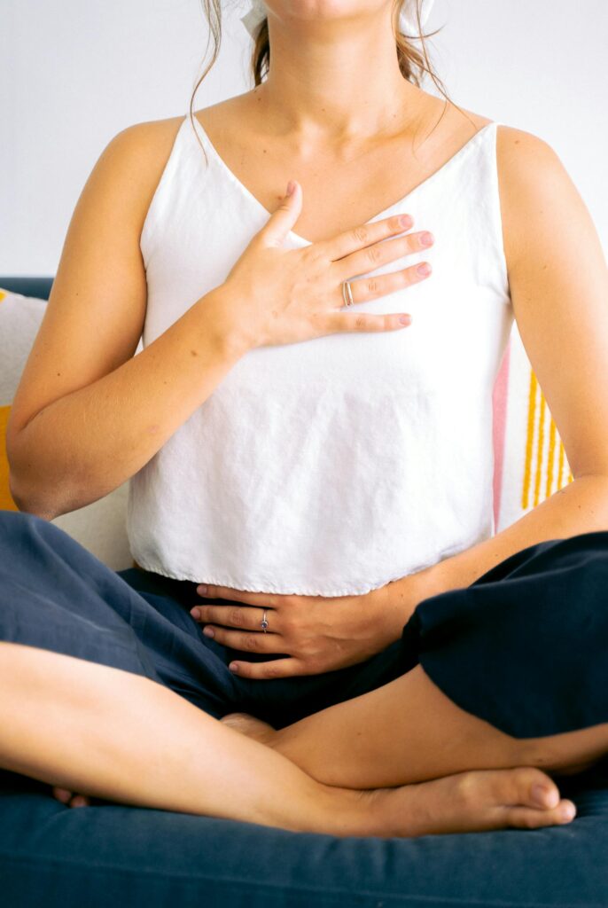 Woman sitting cross-legged in meditation, hands on chest and belly, focusing on breathing and relaxation.