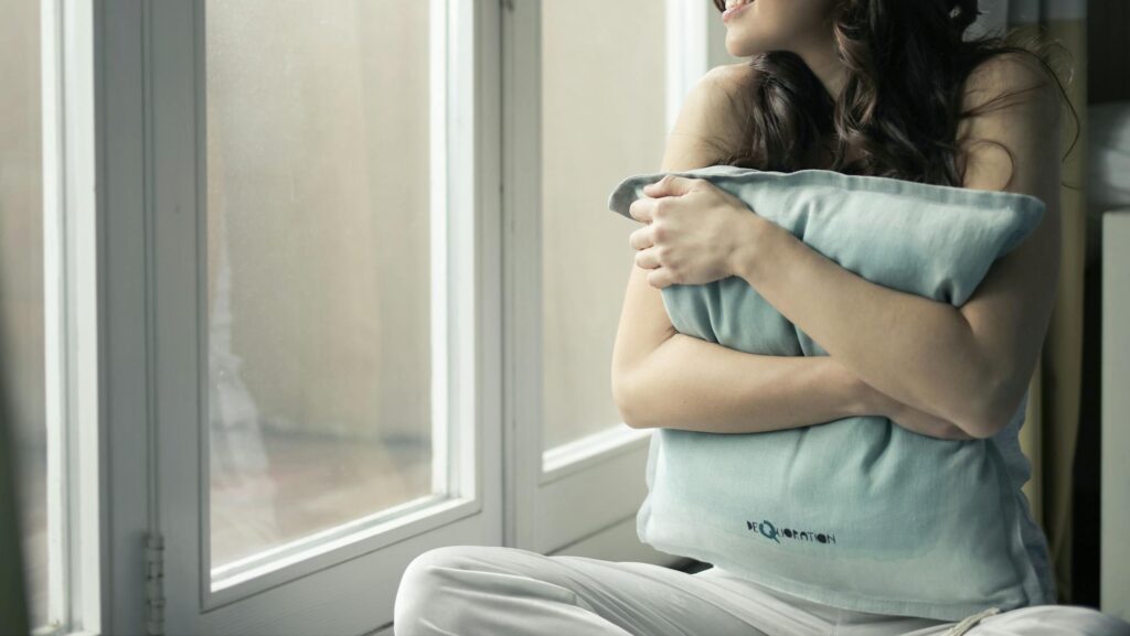 A woman sits by the window, hugging a pillow while smiling warmly during anxiety attack and anxiety disorder healing journey.